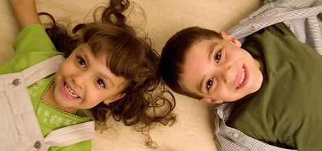 Two smiling children lie on a light-colored surface, with one wearing a green shirt and overalls, and the other in a green shirt with a striped shirt.