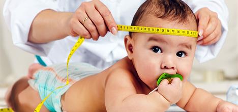 A baby lies on a surface, sucking a pacifier, while a hand measures their head with a tape measure.