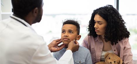 A doctor examines a young boy in a clinic, while his mother watches, holding a teddy bear.
