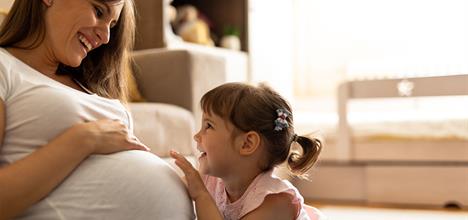 A pregnant woman smiles at a young girl, who is playfully touching her belly, in a cozy indoor setting.