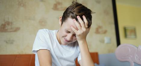 A young boy sits on a bed, holding his head with a pained expression, suggesting discomfort or distress.