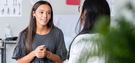 A young woman in a gray shirt is engaged in conversation with another person, likely in a medical or educational setting.